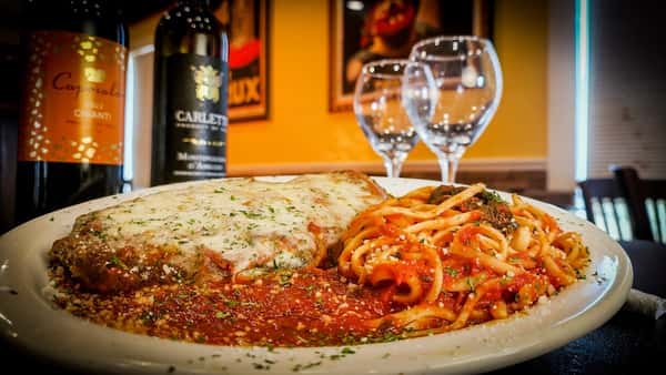 Chicken Parmesan topped with melted cheese, served with a side of spaghetti in marinara sauce, wine bottles and glasses in background.