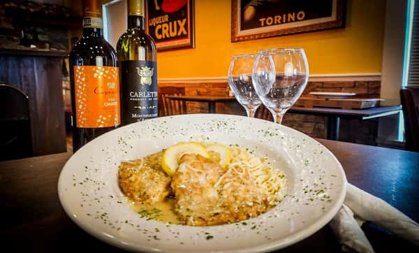 This is a plate of Chicken Piccata served with a side of pasta. The dish features breaded chicken breasts in a lemon butter caper sauce, garnished with lemon slices and sprinkled with herbs and Parmesan. It's elegantly plated with wine and glasses in the background, suggesting a cozy Italian dining setting.