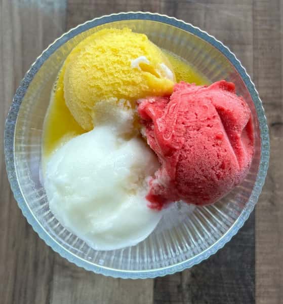 Trio of fruity sorbet scoops in lemon, raspberry, and mango flavors served in a clear glass bowl on a wooden surface.