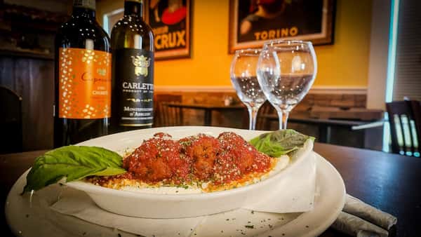 Meatballs in marinara sauce topped with Parmesan, garnished with basil, served beside two wine bottles and glasses.