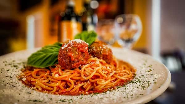 Spaghetti topped with marinara, two meatballs, grated cheese, and basil leaves on a white plate in a cozy restaurant setting.