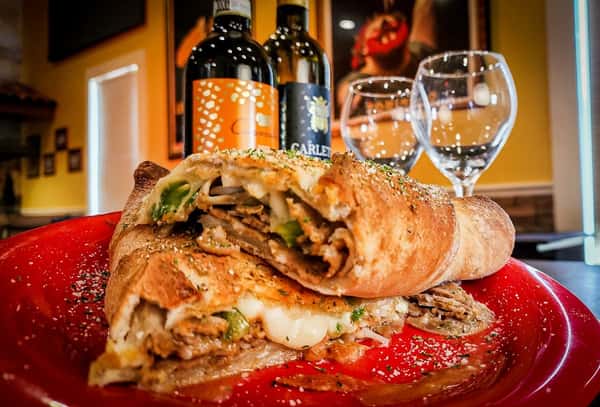 A hearty calzone stuffed with sautéed green peppers, onions, seasoned meat, and melted cheese, served on a vibrant red plate with a sprinkle of herbs and parmesan. Wine bottles and glasses add a cozy Italian restaurant ambiance in the background.
