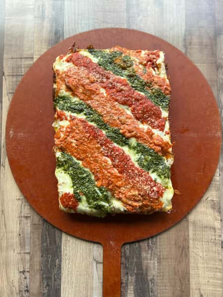 Rectangular lasagna topped with alternating diagonal stripes of red marinara sauce and green pesto on a wooden pizza peel.