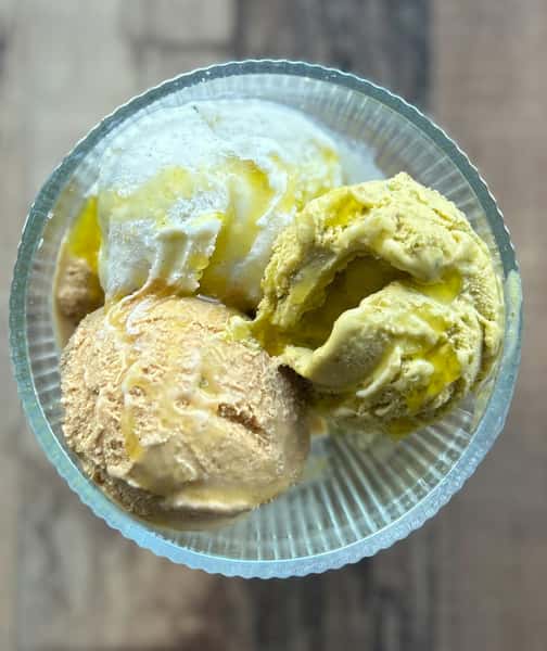 Three scoops of creamy gelato in vanilla, pistachio, and caramel flavors, drizzled with olive oil in a clear dessert bowl.