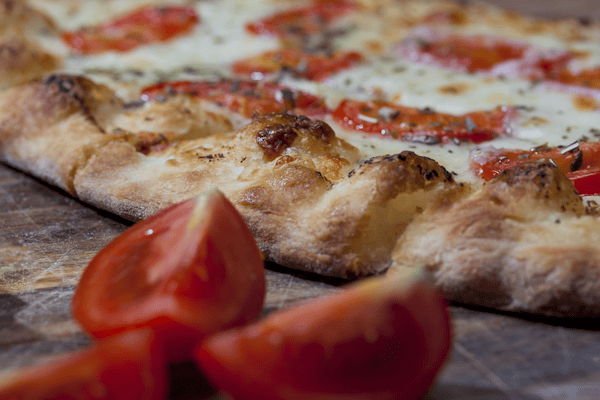Close-up of a crispy margherita pizza with tomato slices, melted mozzarella, and herbs on a rustic wooden surface.