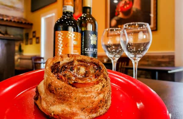 Golden-baked Italian pinwheel roll with meats and vegetables, served on a red plate with wine bottles and glasses in the background.