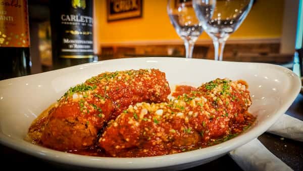 Italian-style meatballs in marinara sauce, topped with Parmesan and parsley, served in a white bowl with wine bottles and glasses behind.