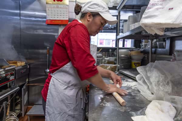 Chinese chef rolling out fresh dough by hand in a flour-dusted kitchen, preparing traditional dumplings or noodles in an authentic restaurant setting.