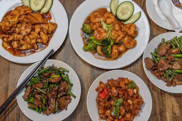 Assorted Chinese stir-fry dishes served on white plates, including spicy chicken with chili sauce, shrimp and broccoli in garlic brown sauce, Kung Pao chicken with bell peppers and peanuts, beef with scallions, and beef with green beans, all presented on a rustic wooden table.