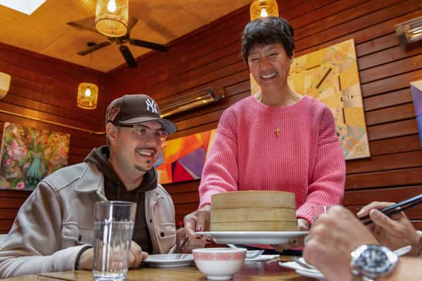 Smiling server presenting bamboo steamers filled with dim sum to happy customers in a cozy, wood-paneled Chinese restaurant with colorful modern art on the walls.