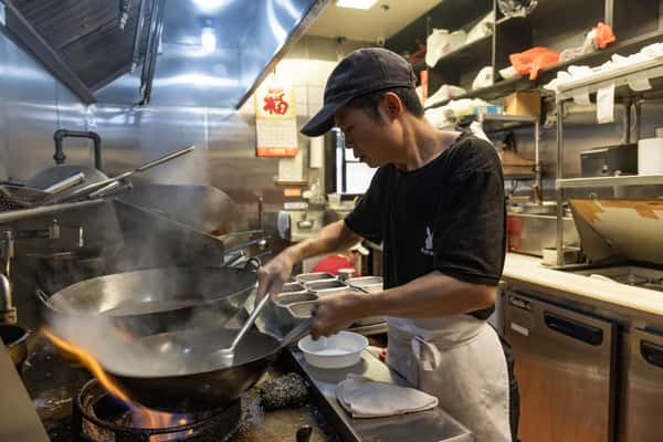 Chinese chef stir-frying over high heat at a commercial wok station, with steam rising in a busy restaurant kitchen during authentic Asian food preparation.