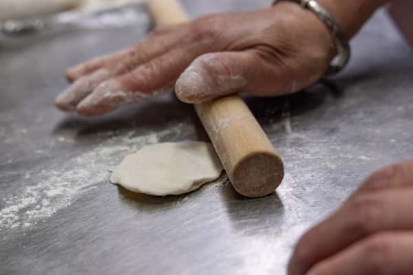 Close-up of hands rolling out dumpling dough on a floured stainless steel surface, showcasing traditional Chinese culinary preparation with a wooden rolling pin.