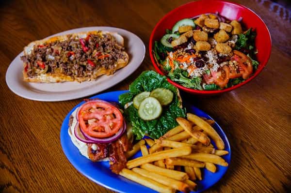 A philly cheese steak, a side salad with croutons, and an open faced burger with grilled bacon, pickles, onions, tomatoes and side fries