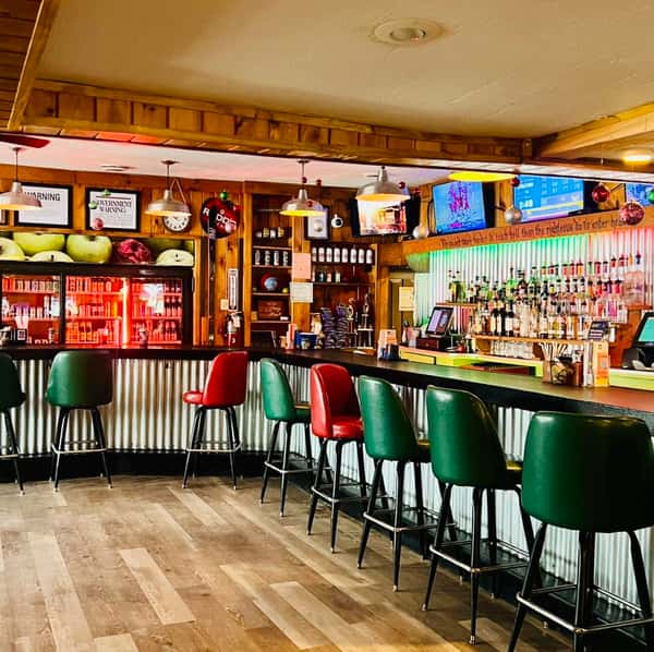 Cozy rustic bar interior with wood-paneled walls, corrugated metal bar front, and red and green vinyl stools, featuring a fully stocked liquor selection, draft beer taps, glowing beverage cooler, and mounted TVs—classic Northwoods tavern ambiance perfect for casual dining and cocktails.