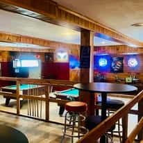 Cozy loft-style seating area overlooking pool tables with wooden railings, neon beer signs, and mounted TVs—classic backwoods bar setting for billiards, drinks, and live sports.