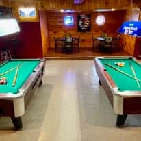 Two green felt pool tables inside a rustic wood-paneled bar with neon beer signs and mounted TVs—classic backwoods tavern game room perfect for billiards, drinks, and casual nightlife.