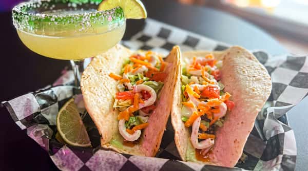 Two tacos with shredded lettuce, cheese, and a sauce, next to a margarita with a lime garnish.