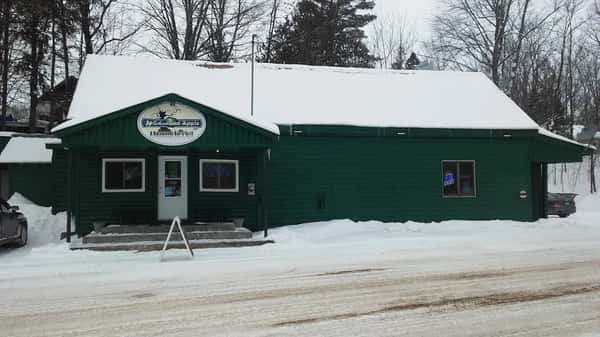 Rustic green roadside restaurant building with snow-covered roof and surrounding winter landscape, featuring a welcoming entrance and cozy Northwoods dining atmosphere.