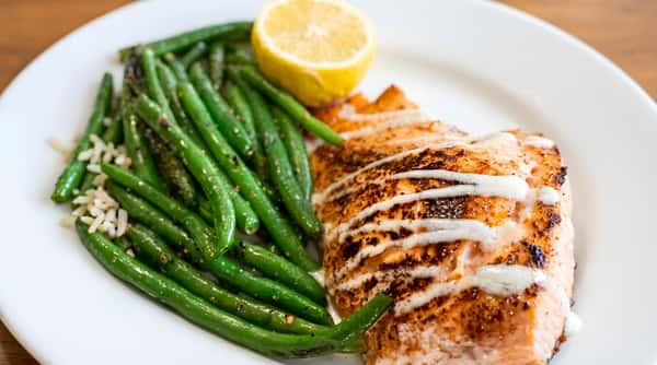 Pan-seared salmon with white drizzle, green beans, and a lemon half on a white plate.