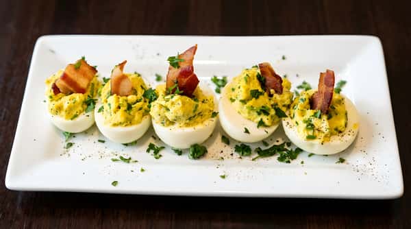 Four deviled eggs on a white rectangular plate, topped with avocado, crispy bacon, and parsley.