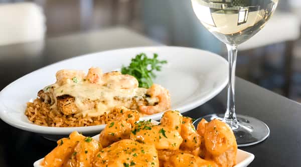 A dish of creamy shrimp over rice and a bowl of fried shrimp with orange sauce, beside a glass of white wine.