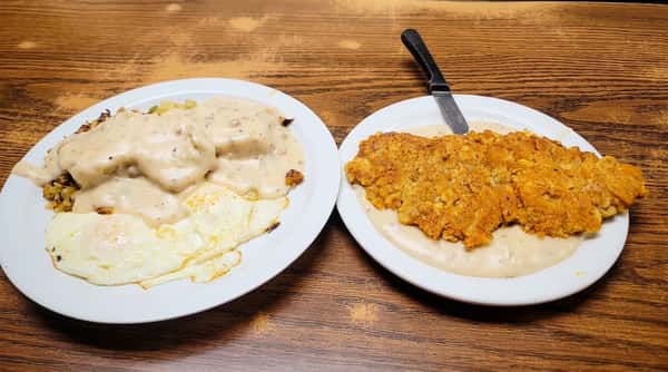 Two plates on a wooden table, one with biscuits and gravy with fried eggs, the other with chicken fried steak.