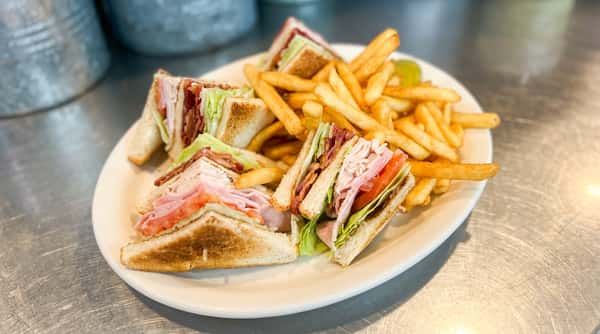 Club sandwich with turkey, ham, bacon, lettuce, tomato, and fries on a white plate with a pickle.