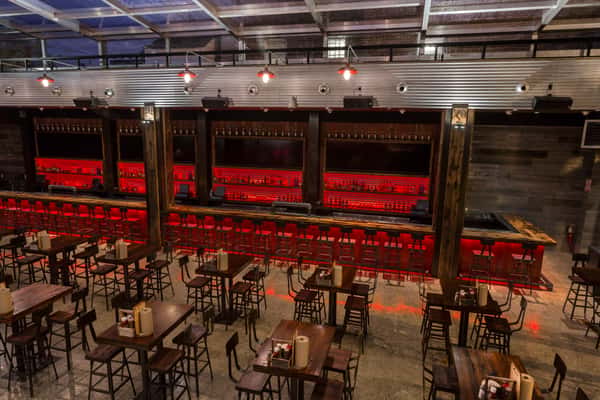 Modern bar interior with high wooden tables and stools, red backlit shelving behind the bar, and a metal ceiling with spotlight fixtures.