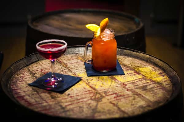 Two cocktails, one red in a sugar-rimmed coupe glass and one orange with orange garnish in a mason jar, on a rustic barrel table.