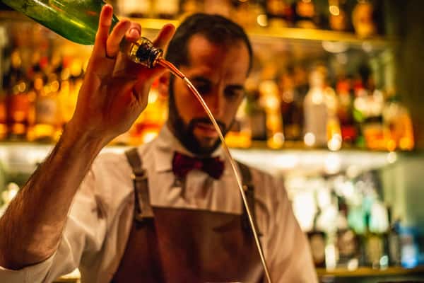 A bartender in formal attire pours a drink with precision behind a warmly lit bar stocked with a variety of liquor bottles.