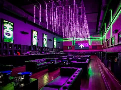 Trendy nightclub with purple and green neon lighting, tufted lounge seating, and digital art displays on the walls.