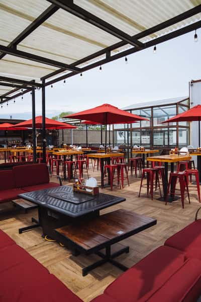 Outdoor rooftop dining space with red umbrellas, high-top tables, and wooden floors under a partially covered pergola.