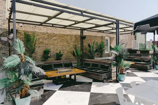 Modern rooftop patio with black-and-white checkered flooring, tropical plants, shaded seating, and industrial-style pergola.