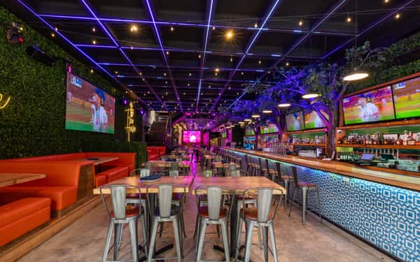 Modern sports bar with neon ceiling lights, red booths, TV screens showing baseball, and a long bar lined with metal stools.