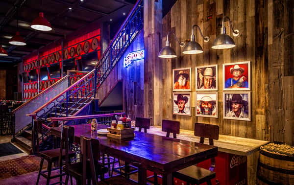 Rustic bar interior with wood-paneled walls, cowboy portraits under metal lamps, and a staircase with red barrel decor and neon signs.