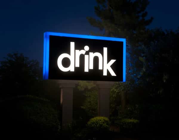 Illuminated outdoor sign reading "drink" in white lowercase letters against a black background with a blue border at night.
