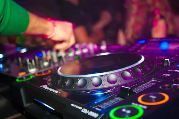 Close-up of a DJ mixing music on a Pioneer CDJ-2000 with colorful lights and a lively nightclub background.