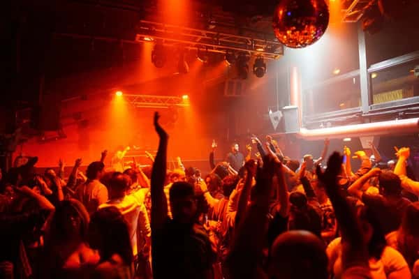 Crowded nightclub dance floor under orange lights with a disco ball and raised hands in the air.
