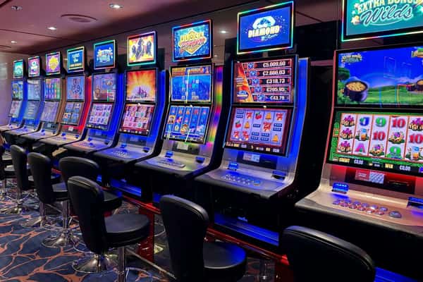 Row of colorful slot machines with flashing screens in a modern casino, empty chairs lined up in front.