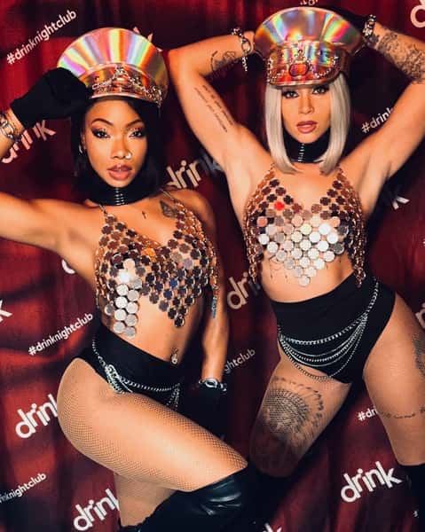 Two performers in holographic hats, metallic sequin tops, fishnet tights, and chains posing at Drink Nightclub.