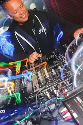 A smiling DJ wearing a "drink" hoodie mixes tracks at a club, surrounded by colorful light trails and professional audio equipment.