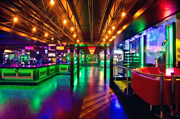 Vibrant nightclub interior with green-lit bar, colorful lights, string ceiling bulbs, and a spiral staircase near a red booth.