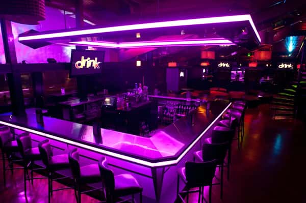 Modern nightclub bar with glowing purple neon lights, high chairs, and sleek interior design under ambient lighting.