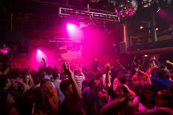 People dancing in a packed nightclub under pink stage lights with a disco ball overhead.