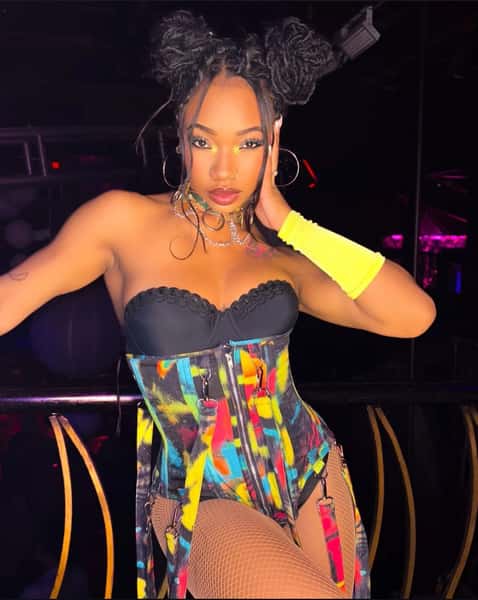 Performer in a colorful corset with black lace top, fishnet tights, neon yellow arm sleeve, hoop earrings, and space bun hairstyle.
