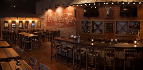 Rustic bar interior with wooden tables and chairs. A vintage Coca-Cola ad painted on a brick wall adds charm. "$2 PBR All day everyday specials".