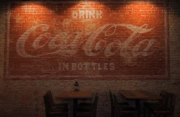 Rustic bar interior with wooden tables and chairs. A vintage Coca-Cola ad is painted on the brick wall. "Drink Coca-Cola in bottles."