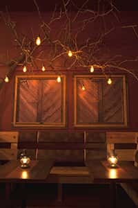 Cozy restaurant interior with rustic wooden tables and chairs. Two vintage framed panels hang behind a booth, illuminated by a decorative tree branch light fixture.