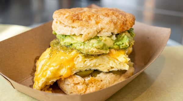 Biscuit sandwich with scrambled eggs, melted cheese, green avocado mash, and a green sauce, in a brown paper food tray.