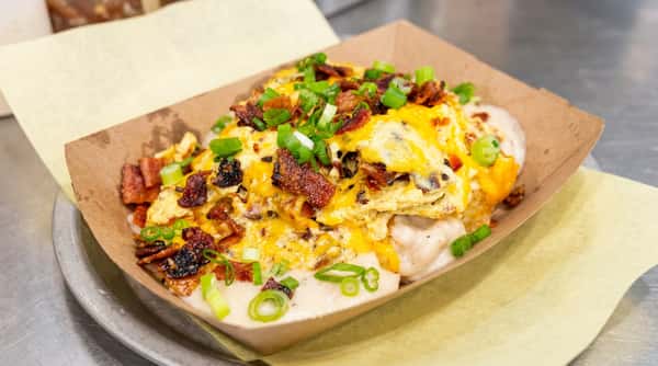 Breakfast bowl with scrambled eggs, melted cheese, bacon bits, and chopped green onions in brown paper tray.